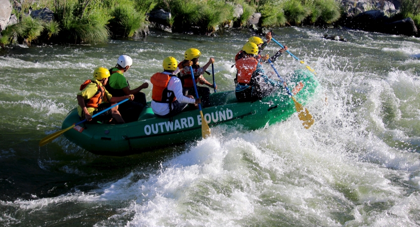 a group of outward bound students wearing helmets and life jackets paddle a raft through whitewater